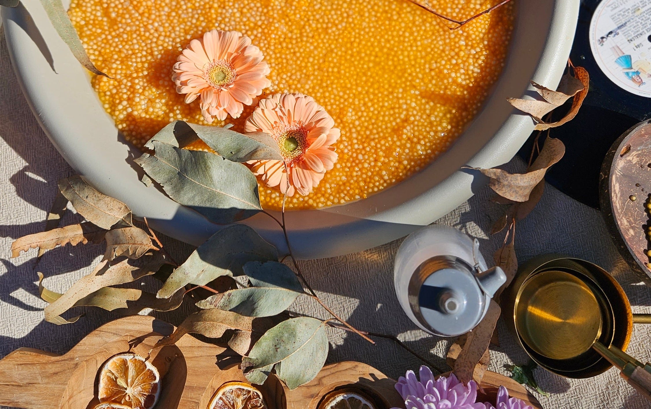 A styled overhead flat-lay of a large white bowl filled with golden yellow grains or seeds, decorated with dried leaves and pink flowers, surrounded by small wooden bowls, dried citrus slices, purple flowers, and a teapot on a light fabric surface.